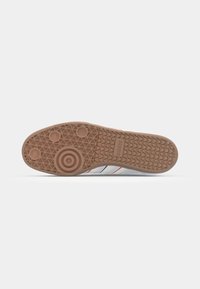 Sneaker gris clair avec semelle en caoutchouc naturel, présentant des rayures bleues et rouges. La semelle a un motif texturé avec des détails circulaires et antidérapants.