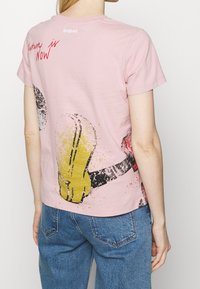 Una persona con el cabello largo, vestida con una camiseta rosa que presenta coloridos diseños gráficos y texto, está de espaldas al espectador.