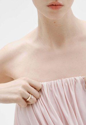 Primer plano de una persona sosteniendo una tela fruncida rosa cerca de la clavícula, con un anillo dorado con una única piedra preciosa clara.