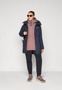 Naine, kes kannab päikeseprille, tumesinist jakki, mauve värvi tõmblukuga kihilise sviitriga, musti pükse, mauve värvi matkajalatsid ning kannab musta seljakoti rihma.