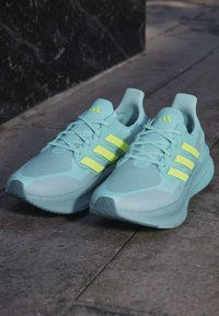 Ein Paar hellblaue Laufschuhe mit neonfarbenen gelben Streifen und Schnürsenkeln, die auf einem steinernen Pflaster neben einer schwarzen Wand platziert sind.