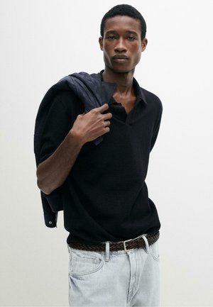 Giovane uomo con maglione nero e jeans chiari, che tiene una giacca scura sulla spalla davanti a uno sfondo chiaro uniforme.