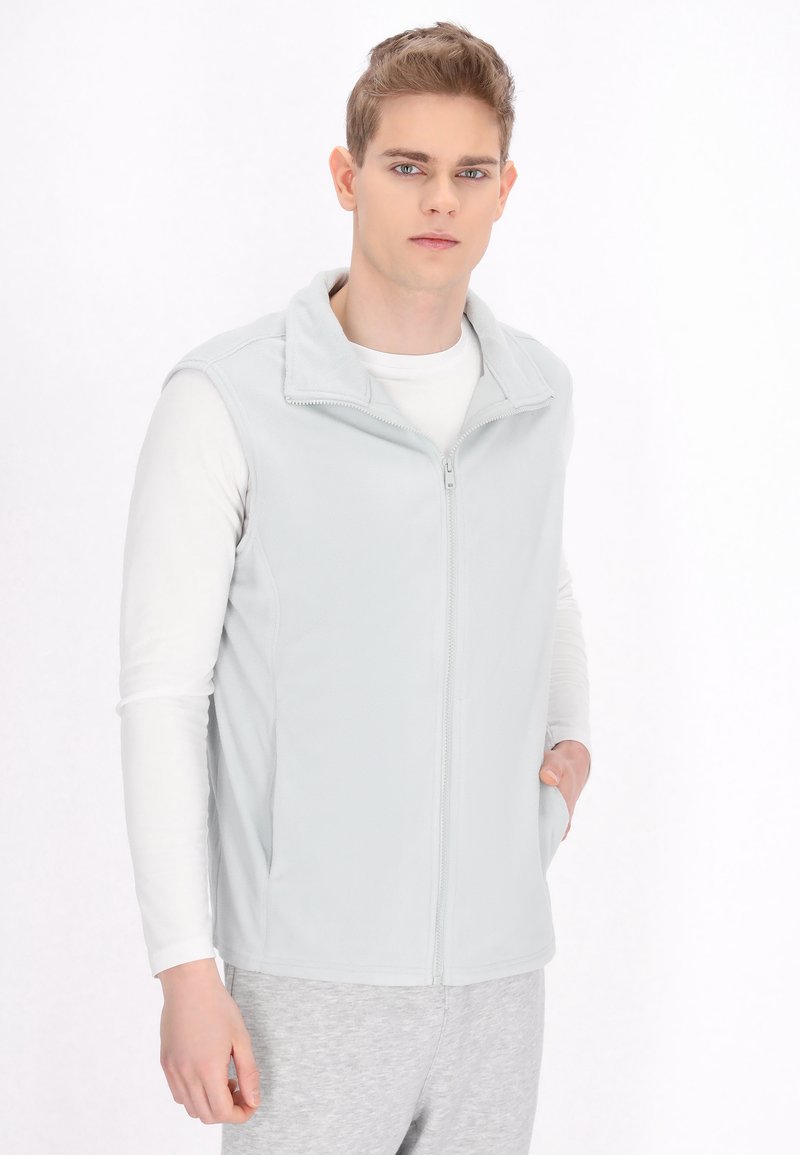 Ljusgrå fleeceväst med zippered front, hög krage och sidfickor, bärs över en vit långärmad skjorta och grå byxor.