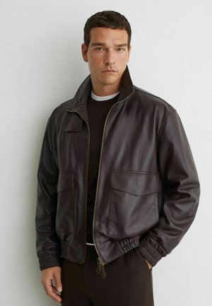 Mann trägt eine dunkelbraune Lederjacke mit großen Vorderentaschen und elastischen Bündchen, steht an einer hellen Wand.