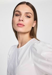 Biała bluzka z bufkami i subtelnym haftem kwiatowym. Gładka tekstura, okrągły dekolt i luźny krój.