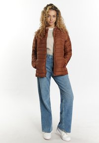 Brun vadderad jacka med hög krage, dragkedjestängning och sidofickor, kombinerad med ljusblå vida jeans och vita sneakers.