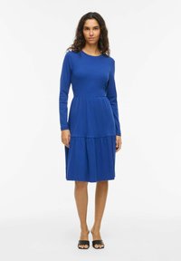Modré šaty s dlouhým rukávem, s přiléhavým vrškem a nabíranou, vrstvenou sukní. Vyrobené z měkké, pružné látky, s kulatým výstřihem a volným střihem.