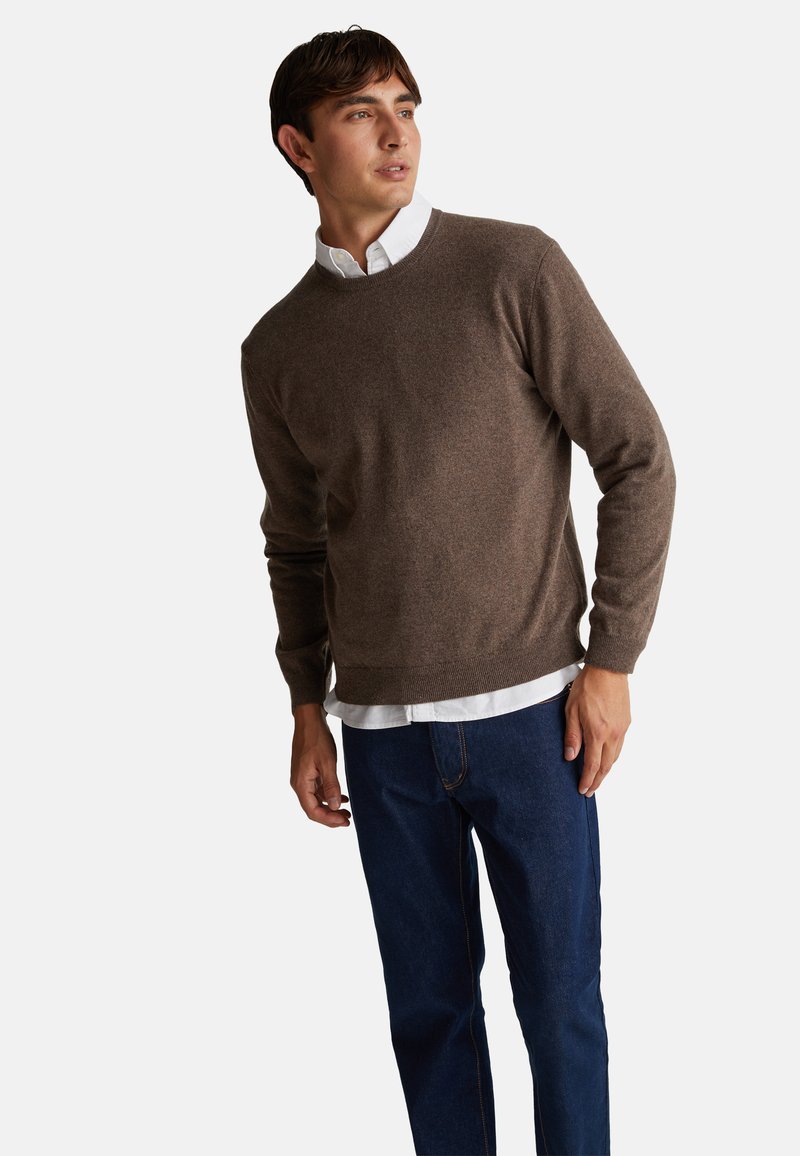 Maglione marrone a maglia con scollo rotondo, abbinato a una camicia bianca con colletto sotto. Indossato con jeans blu, che mostrano un look casual.