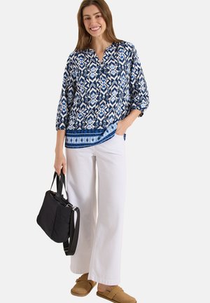 Smilende kvinde iført bluse med blåt mønster, brede hvide bukser, lysebrune slip-on sko, der holder en sort håndtaske med den ene hånd i lommen.