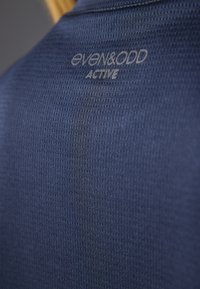 Blå atletisk skjorte med en tekstureret stofmønster og synlig branding "even&odd ACTIVE" i lys grå på den øverste del af ryggen.