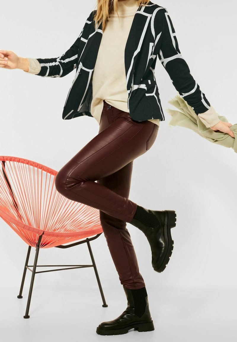 Femme portant un blazer à motif noir et blanc, un pull beige, un pantalon en cuir bordeaux et des bottes noires, en équilibre sur une jambe près d'une chaise rouge.