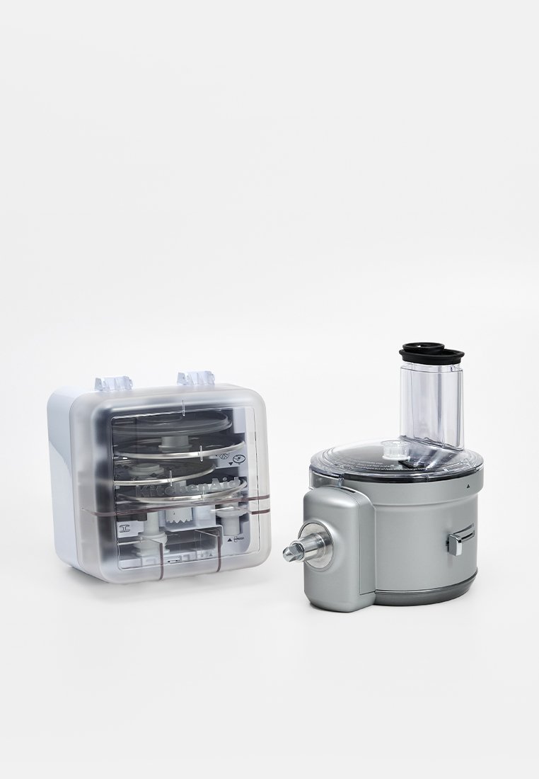 Robot culinaire avec un corps argenté et un récipient de stockage transparent pour les accessoires. Présente une large ouverture d'alimentation et divers disques de découpe visibles à l'intérieur.