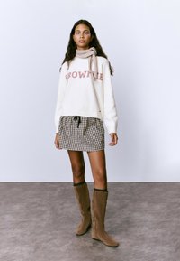 Suéter blanco con "BROWNIE" en rosa, combinado con una falda a cuadros y botas marrones hasta la rodilla. El suéter tiene una textura suave y un corte relajado.