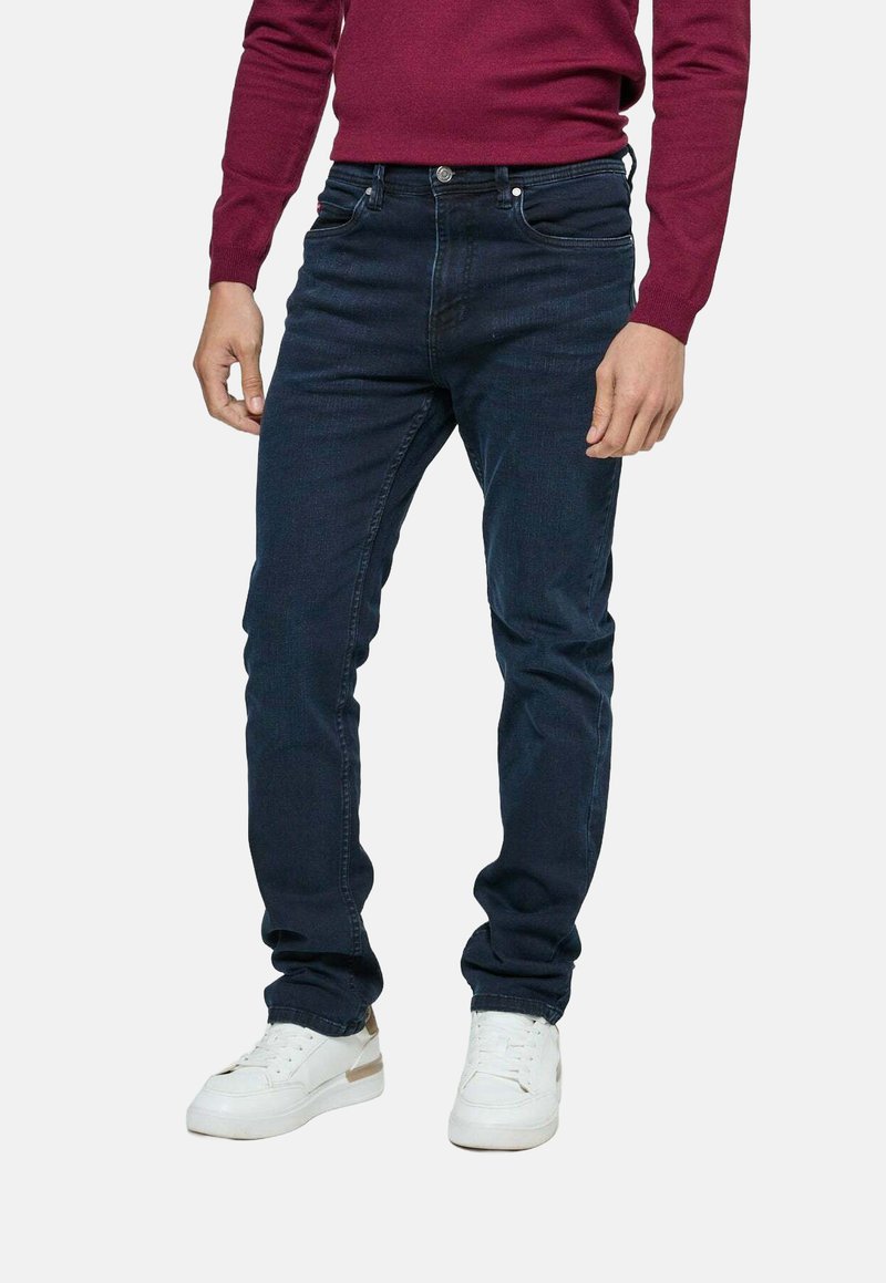 Homme portant un jean bleu foncé, des baskets blanches et un pull à manches longues bordeaux, se tenant sur un fond gris uni.