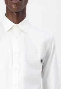 Vit långärm skjorta med klassisk krage, med knäppning och ett slätt, lätt tyg. Snygga sömnadsdetaljer är synliga.