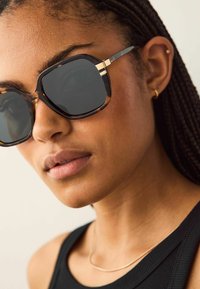 Braune Schildpatt-Sonnenbrille mit großen quadratischen Fassungen und dunklen Gläsern. Dünne goldene Akzente in der Nähe der Schläfen verleihen Details.