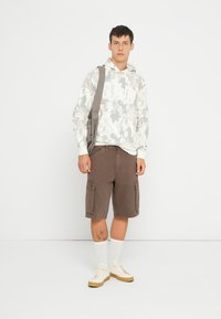Felpa camo grigia chiara con tasca frontale, pantaloni corti cargo marroni con tasche laterali, calze bianche e sneakers beige. Design semplice e informale.
