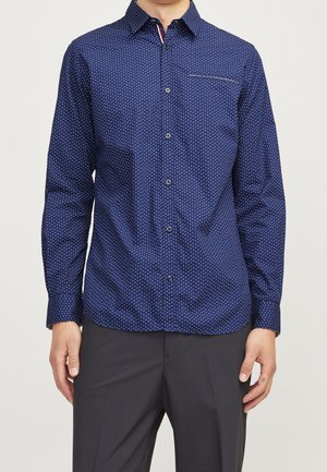 Man draagt een nauwsluitend navyblauw overhemd met patroon en lange mouwen, gecombineerd met een donkere broek, tegen een effen achtergrond.