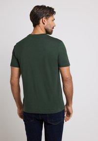 Camiseta de manga corta verde oscuro hecha de algodón, presenta un cuello redondo y un diseño liso con costuras cuidadas en el dobladillo y las mangas.
