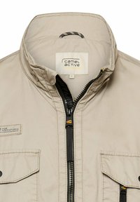 Beige Jacke mit Reißverschluss und Stehkragen, zwei Brusttaschen mit Knopflaschen und einem "camel active"-Label innen im Kragen.