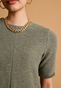 Oliwkowa, teksturowana bluzka z krótkim rękawem i okrągłym dekoltem, z widocznym detalem szwu, zestawiona z masywnym złotym naszyjnikiem.