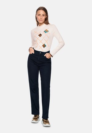Langärmliges cremefarbenes Oberteil mit buntem geometrischen Muster; dunkelblaue gerade Jeans; Turnschuhe mit Leopardenmuster und cremefarben.