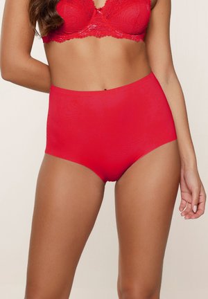 Vrouw in een bijpassende rode kant bhs en hoge naadloze slip, van halverwege de romp tot halverwege de bovenbenen, tegen een neutrale achtergrond.