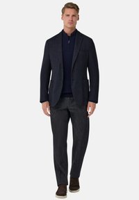 Námořnicky modré texturované sako se dvěma předními kapsami, spojené s tmavým svetrem na zip a tmavými vzorovanými kalhotami, doplněné hnědými botami.
