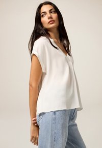 Blusa blanca de manga corta con diseño de escote en V, hecha de una tela ligera, combinada con jeans de mezclilla azul claro que cuentan con detalles decorativos.