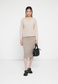 Femme portant un cardigan beige boutonné, une jupe midi en maille assortie, des chaussures Mary Jane noires et tenant un petit sac à main noir.