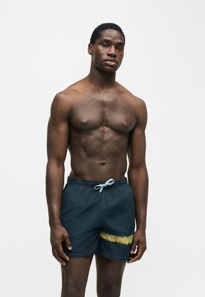 Homme musclé à la peau foncée portant un short de bain bleu marine avec un détail de coup de pinceau jaune, debout devant un fond clair uni.