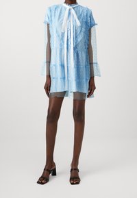 Ljusblå genomskinlig klänning med volangdetaljer och knäppning fram, med långa genomskinliga ärmar och en vit snodd vid kragen. Svarta klacksandaler.