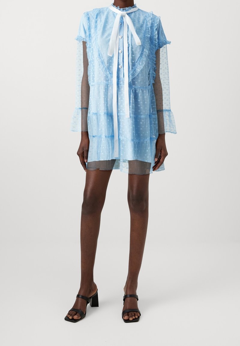 Ljusblå genomskinlig klänning med volangdetaljer och knäppning fram, med långa genomskinliga ärmar och en vit snodd vid kragen. Svarta klacksandaler.