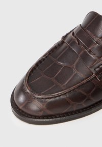 Bruine leren loafers met krokodillenstructuur, moc toe stiksels en een decoratieve band over de bovenkant, weergegeven op een witte achtergrond.