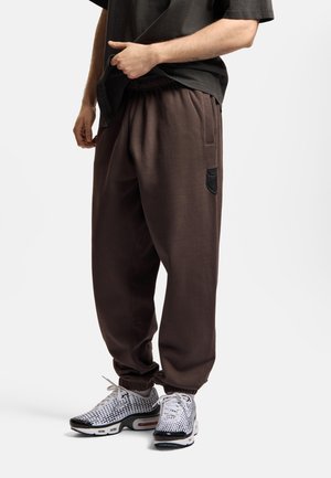 Mężczyzna ubrany w ciemnobrązowe spodnie dresowe z elastycznymi ściągaczami i białe-czarne wzorzyste sneakersy, trzymający czarną koszulkę.