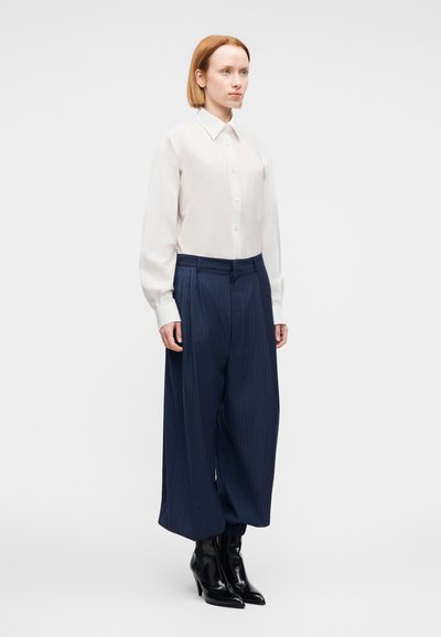 Femeie cu păr scurt roșcat, purtând o cămașă albă cu nasturi, pantaloni largi bleumarin cu dungi fine și cizme negre cu toc mediu, stând pe un fundal alb.