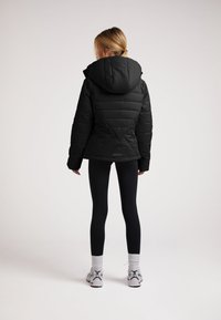 Giubbotto piumino nero con cappuccio, caratterizzato da una trapuntatura orizzontale e un design triangolare sul retro. Indossato sopra leggings neri e sneaker.