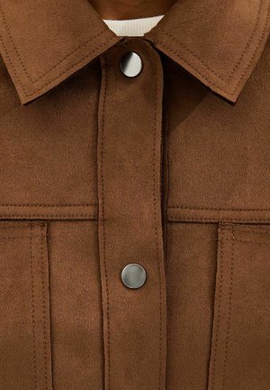 Bruine suède jas met twee zilveren drukknopen, zichtbare stiksels en een brede kraag over een wit shirt.