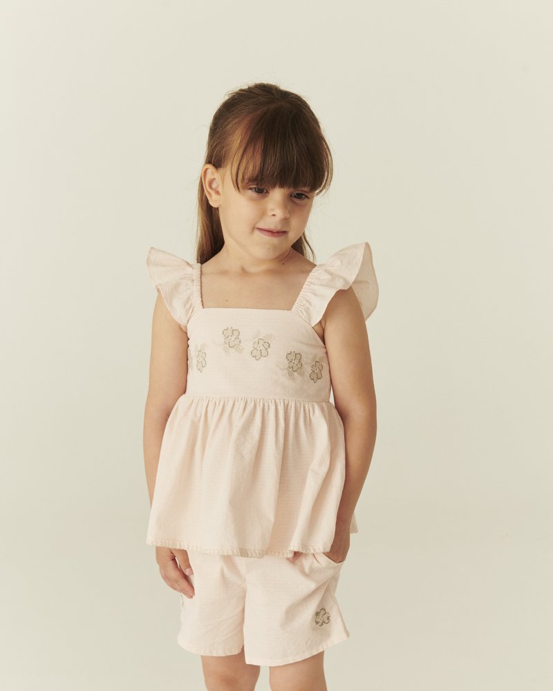 Mladé dievča s hnedými vlasmi, ktoré má na sebe svetloružový volánikový vrchný diel bez rukávov a zodpovedajúce šortky s kvetinovou výšivkou na jednofarebnom pozadí.