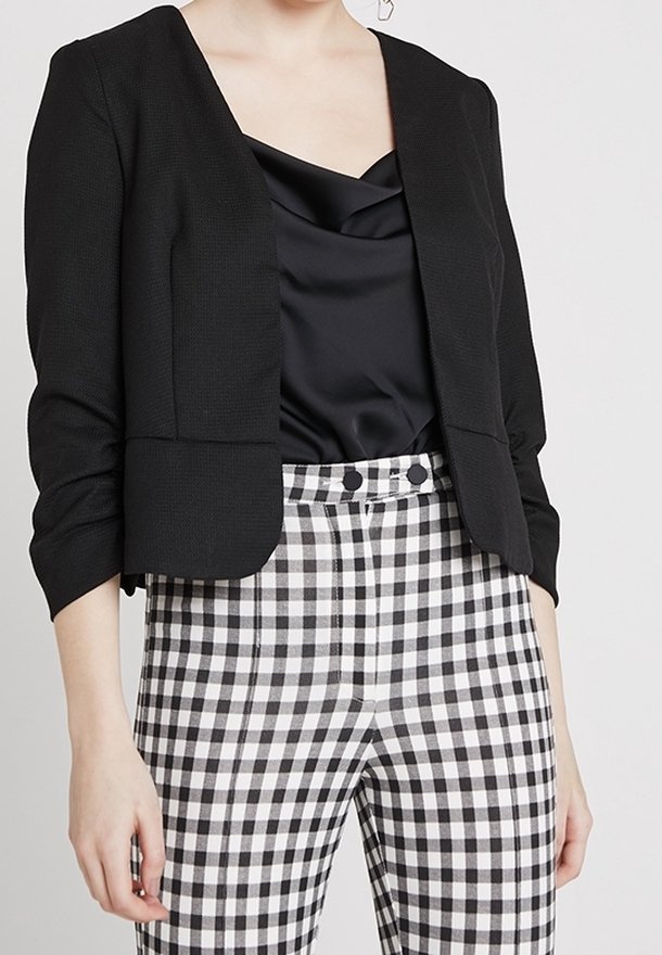 Femme portant un blazer noir court, un top en satin noir et un pantalon taille haute à carreaux noirs et blancs avec des boutons.