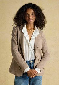 Femme aux cheveux bouclés portant un blazer beige, un chemisier blanc à volants, un collier chaîne en or et un jean bleu, debout les mains jointes.