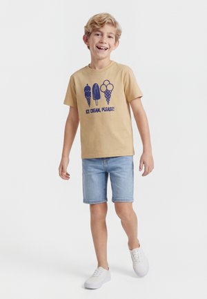 Smiješeći se dječak koji nosi bež majicu s grafikom sladoleda i tekstom "SLADOLED, MOLIM!", traper kratke hlače i bijele tenisice, hoda naprijed.