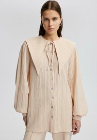 Blusa texturizada beige con un gran cuello estructurado, cierre de lazo y frente de botones. Mangas largas con puños fruncidos. Tela suave.