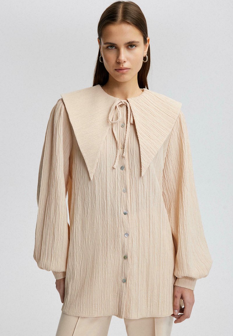 Blusa texturizada beige con un gran cuello estructurado, cierre de lazo y frente de botones. Mangas largas con puños fruncidos. Tela suave.