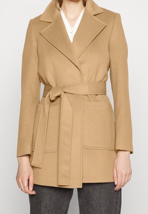 Kvinde iført en kamelfarvet uldfrakke med bælte, notched revers og store frontlommer over mørkegrå bukser.