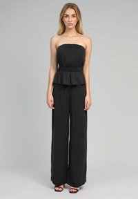 Combinaison noire sans bretelles avec un corsage ajusté et une taille peplum, associée à des pantalons à jambes larges. Tissu doux et chaussures à bride de cheville visibles.