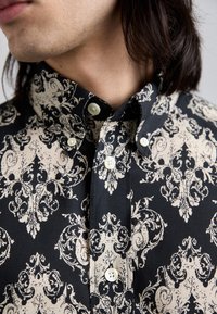 Camicia a fantasia nera e beige con un design floreale decorativo, colletto button-down e bottoni bianchi, realizzata in un tessuto texturizzato.