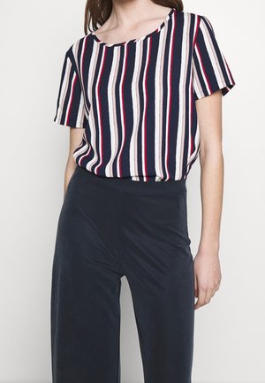 Vrouw die een top met korte mouwen draagt met verticale strepen in marineblauw, wit, beige en rood, gecombineerd met een high-waisted broek in donker marineblauw.