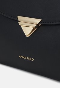 Sac à main en cuir noir avec fermeture triangulaire en métal doré et le nom de la marque "ANNA FIELD" embossé en doré sous la fermeture.