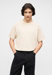 Νεαρός με κοντά κυματιστά σκούρα μαλλιά, που φοράει μπεζ μπλουζάκι με στρογγυλή λαιμόκοψη και μαύρο παντελόνι, στέκεται με τα χέρια στις τσέπες μπροστά σε απλό φόντο.